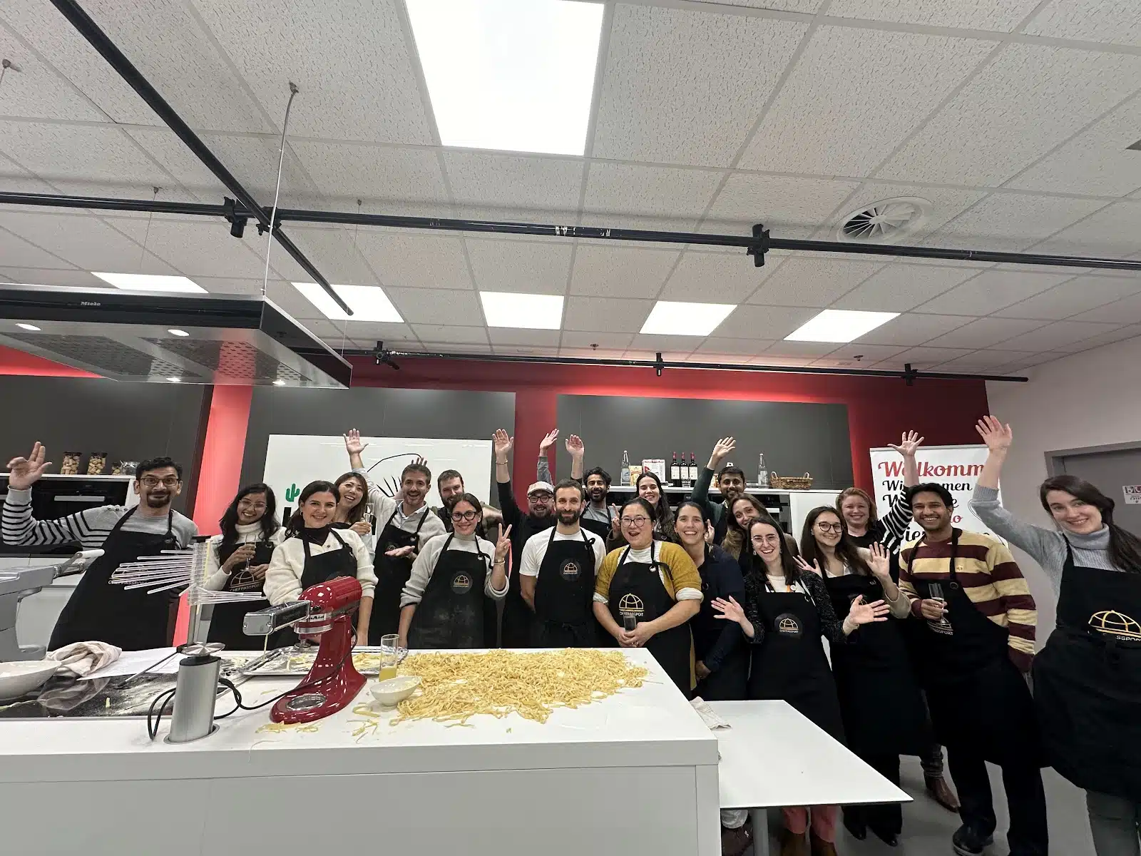 A large group of smiling people in aprons wave at the camera during a culinary team building event, with fresh pasta on a counter.